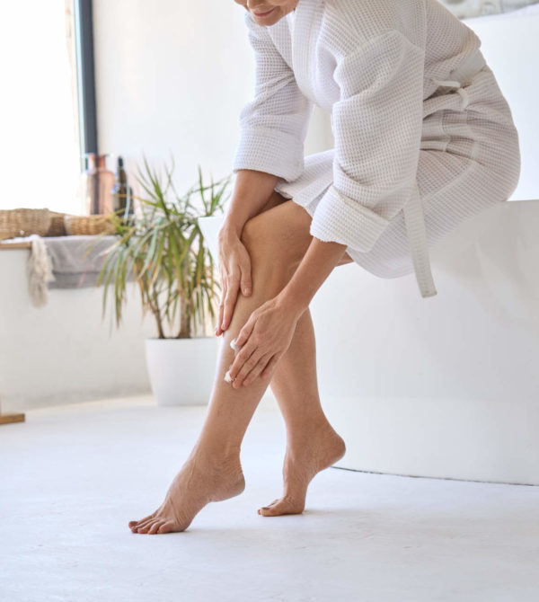 Mid age adult 50s age mature woman applying varicose prevention treatment cream massaging legs sitting on bathtub wearing white bathrobe. Feminine health care after menopause concept.
