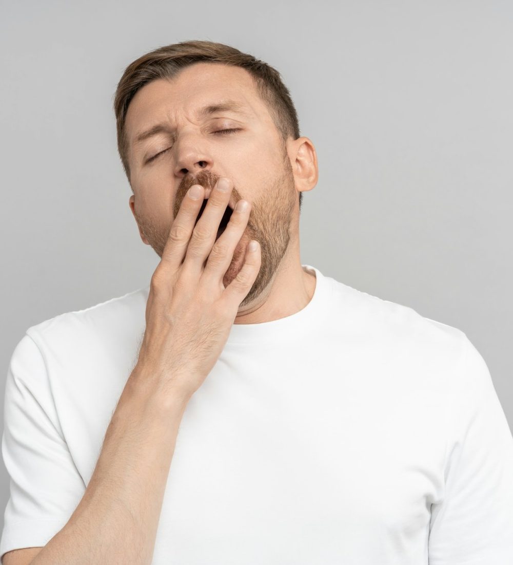 Sleepy yawning man with closed eyes opened mouth covering hand on gray background.